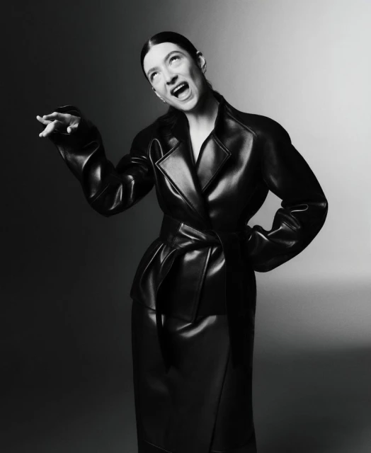 Black and white studio shot of a woman in a glossy black trench coat and matching skirt with her mouth open and arm extended.