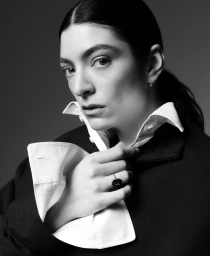 Portrait of singer Lorde adjusting her white collared shirt under a dark jacket against a plain grey studio backdrop.