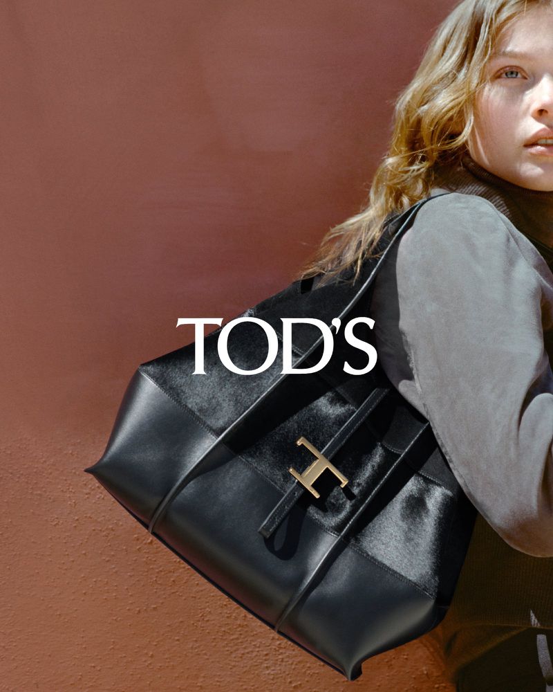 Fashion shot featuring a Tod's black leather handbag with blonde model; focus on bag against a warm brown studio backdrop with the brand's logo.