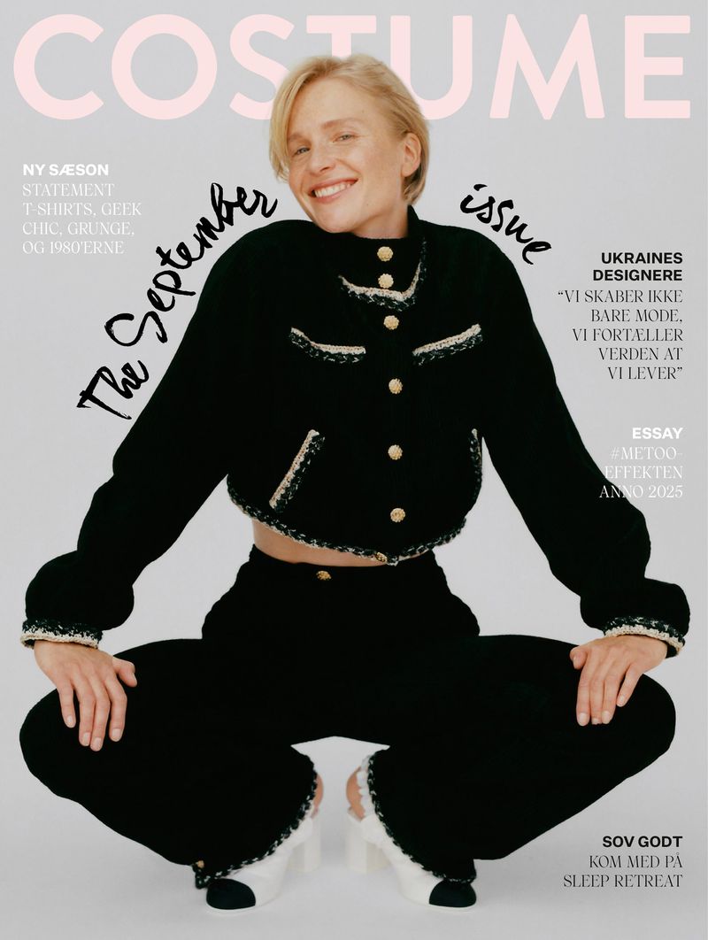 Blonde woman in black tweed jacket and wide-leg pants squats for fashion magazine cover, smiling at the camera in a photo studio setting.