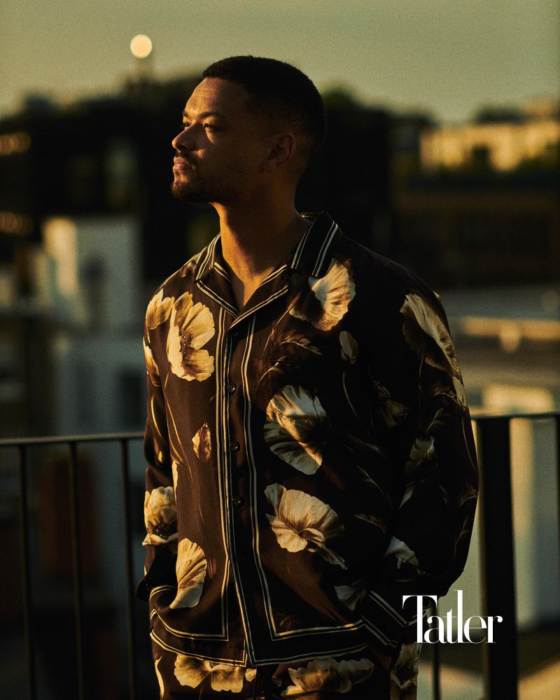Portrait of an adult male in a patterned shirt on a rooftop, gazing to the side, captured at eye-level with a shallow depth of field.