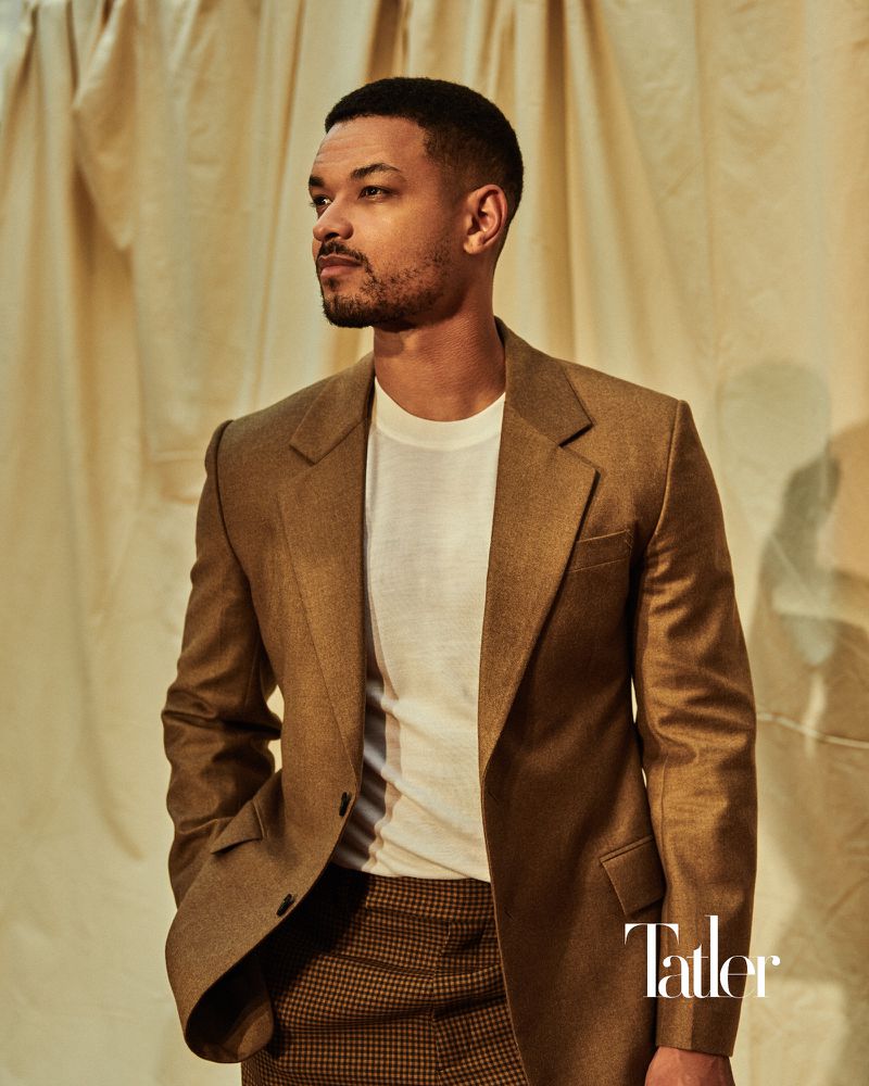 Portrait of a man in a brown suit jacket and patterned pants looking away against a draped cloth background.