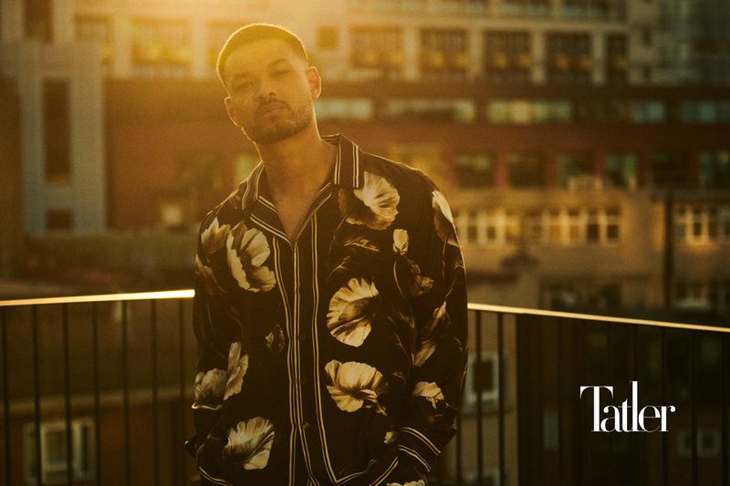 Portrait of adult male in floral shirt standing on a balcony at sunset in front of apartment building with Tatler logo in the corner.