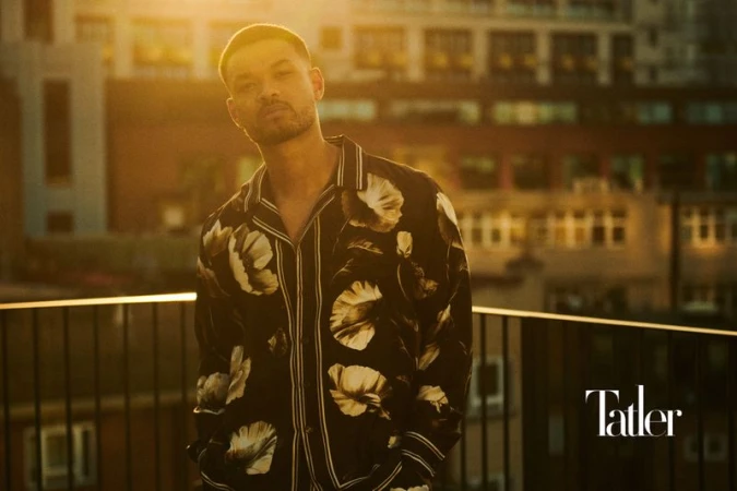 Portrait of adult male in floral shirt standing on a balcony at sunset in front of apartment building with Tatler logo in the corner.
