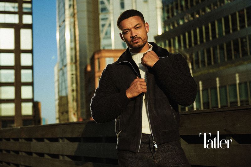 Steven Bartlett wearing a black zip jacket and white high-neck knit standing on an urban rooftop against city buildings.