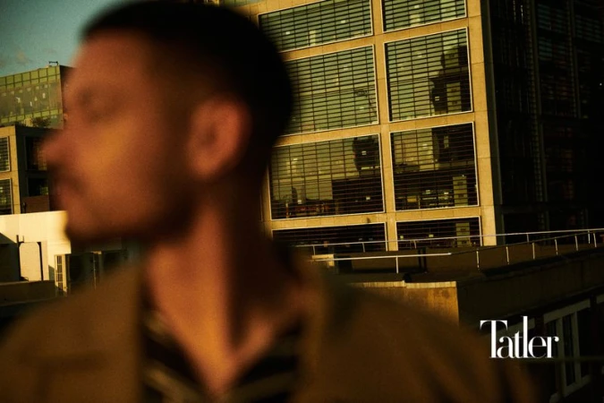 An out-of-focus portrait of a man is shown against a background featuring an urban building with many windows under a golden light.