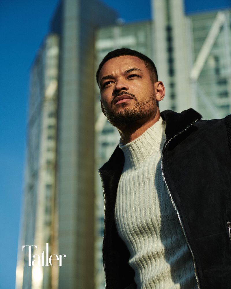 Steven Bartlett wearing a cream ribbed turtleneck and black vest in front of modern skyscrapers against a blue sky.