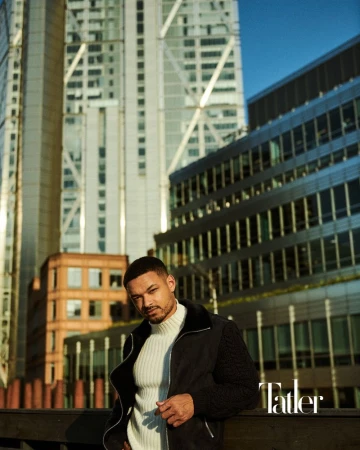 Steven Bartlett wearing a black zip jacket and cream ribbed turtleneck sweater outdoors against urban office skyscrapers.