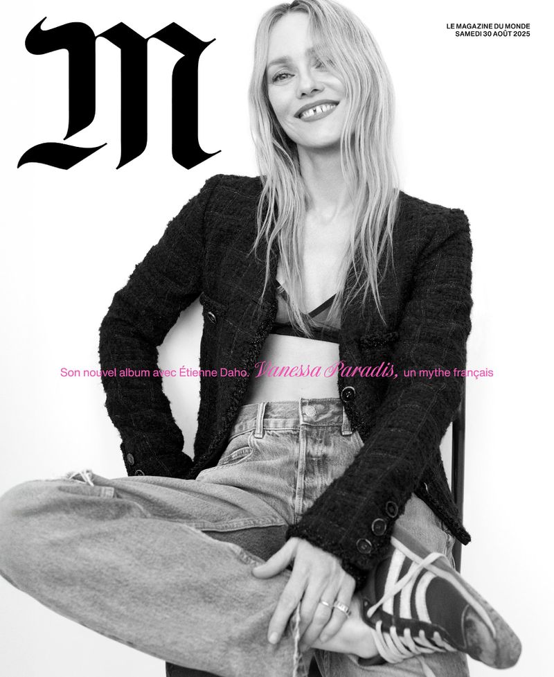 A woman with blonde hair, wearing a dark jacket and jeans, smiles while sitting against a plain white background.