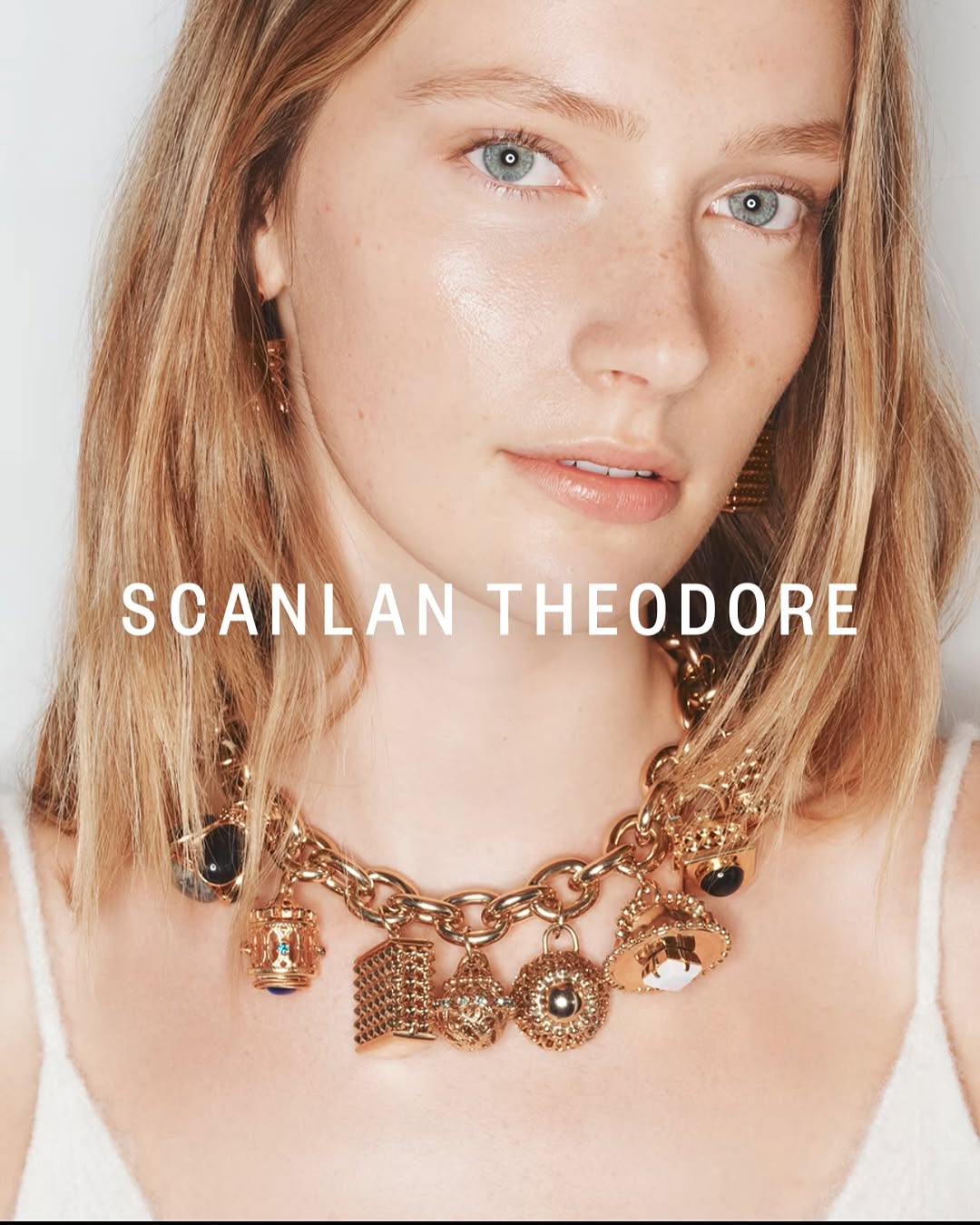 Close-up of a blonde woman wearing a gold chain necklace with several charms. Beauty shot with soft light and a white background.
