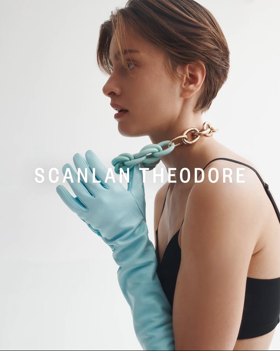 Close-up fashion portrait of a young woman in a black top and blue gloves, wearing a gold and blue necklace against a white background.