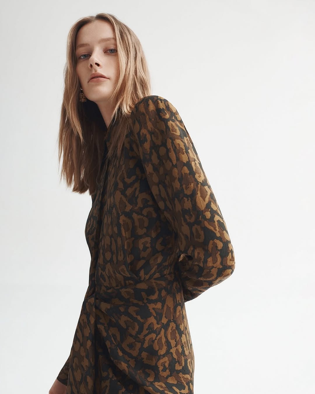 Young woman in brown and black leopard print long-sleeve dress poses in a minimalist studio with soft, diffused lighting.