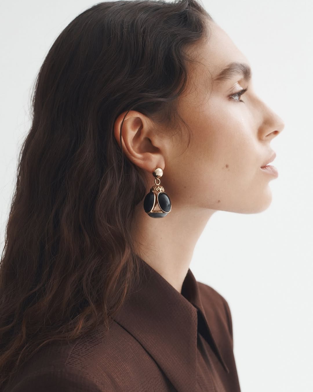 A woman is seen in a profile shot, showcasing dark, wavy hair and a brown collared shirt, complemented by black and gold earrings.