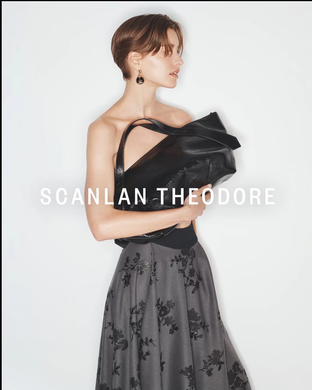 Model with short brown hair wearing a grey floral skirt and holding a large black leather handbag against a white background