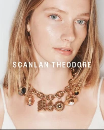 Close-up portrait of a woman wearing a heavy gold charm necklace and a white top against a neutral background.