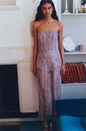 Elegant adult woman with long dark hair poses in a shimmering strapless jumpsuit in a light-filled interior with a fireplace.