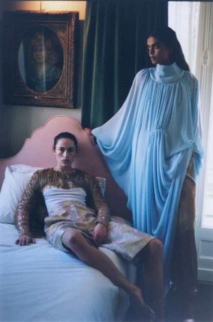 Two fashion models pose indoors, one seated on a bed in a printed dress, the other standing in a blue chiffon gown next to pink headboard.