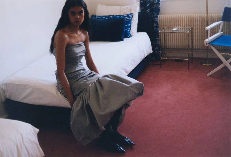 A South Asian woman sits in a silver strapless dress on a bed in a brightly lit room, her gaze focused directly at the viewer.