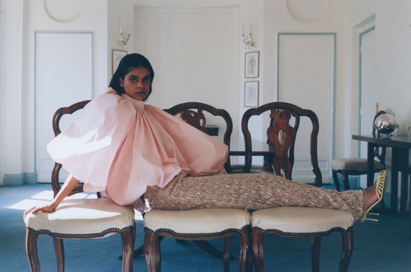 A South Asian woman in a pink top and patterned pants lounges across three ornate chairs in a white-walled room with blue carpet.