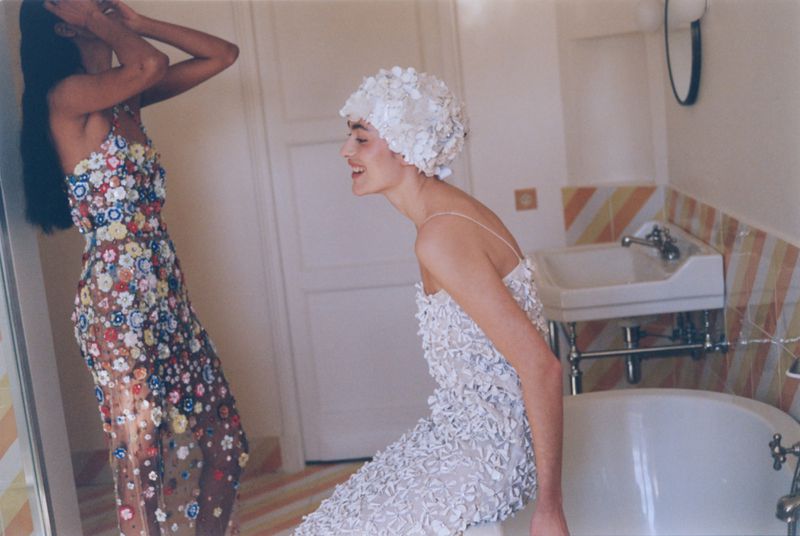 Two women in dresses with floral embellishments pose in a bathroom with a tub and sink, one wearing a white floral swim cap.
