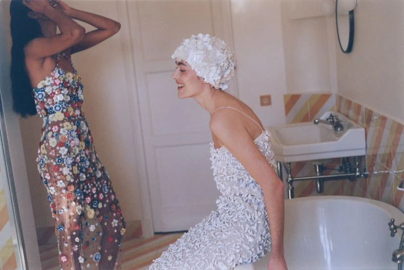 Two women in dresses with floral embellishments pose in a bathroom with a tub and sink, one wearing a white floral swim cap.