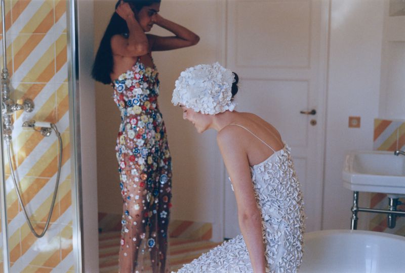 Two women in ornate dresses and hats pose in a bathroom with yellow-striped tiling, white doors, and a vintage bathtub.