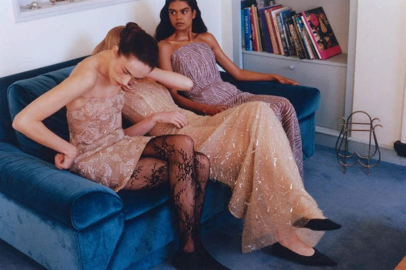 Three women in elegant dresses sitting together on a teal velvet couch, indoor setting, one model is wearing black patterned tights.