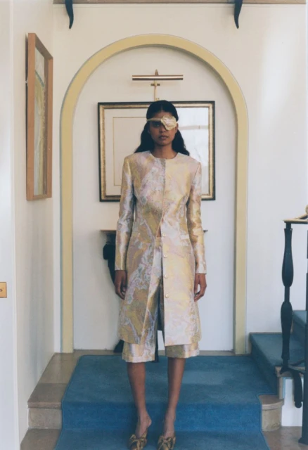 A woman stands indoors, wearing a gold patterned jacket and shorts, with a decorative eye patch, in a building with cream archways.