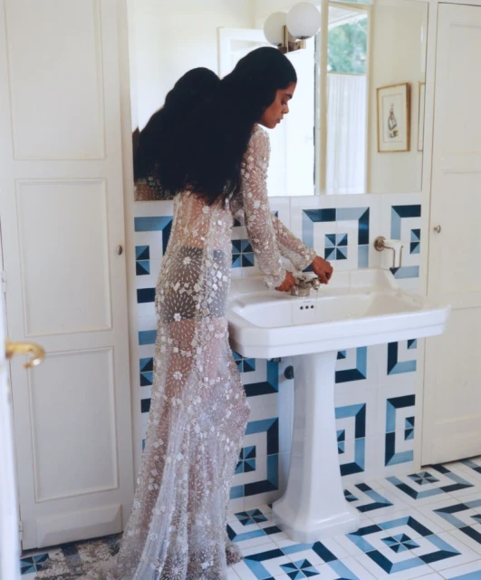 Woman in an embellished dress washes her hands in a white bathroom with blue patterned tile, under modern lighting and a mirror.