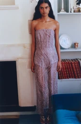 Model wearing a strapless sheer beaded lavender column gown standing beside a fireplace and bookshelf.