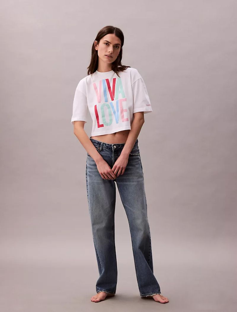 Model Mili Boskovic standing barefoot wearing a white Viva Love cropped t-shirt and blue denim jeans against a grey studio backdrop.