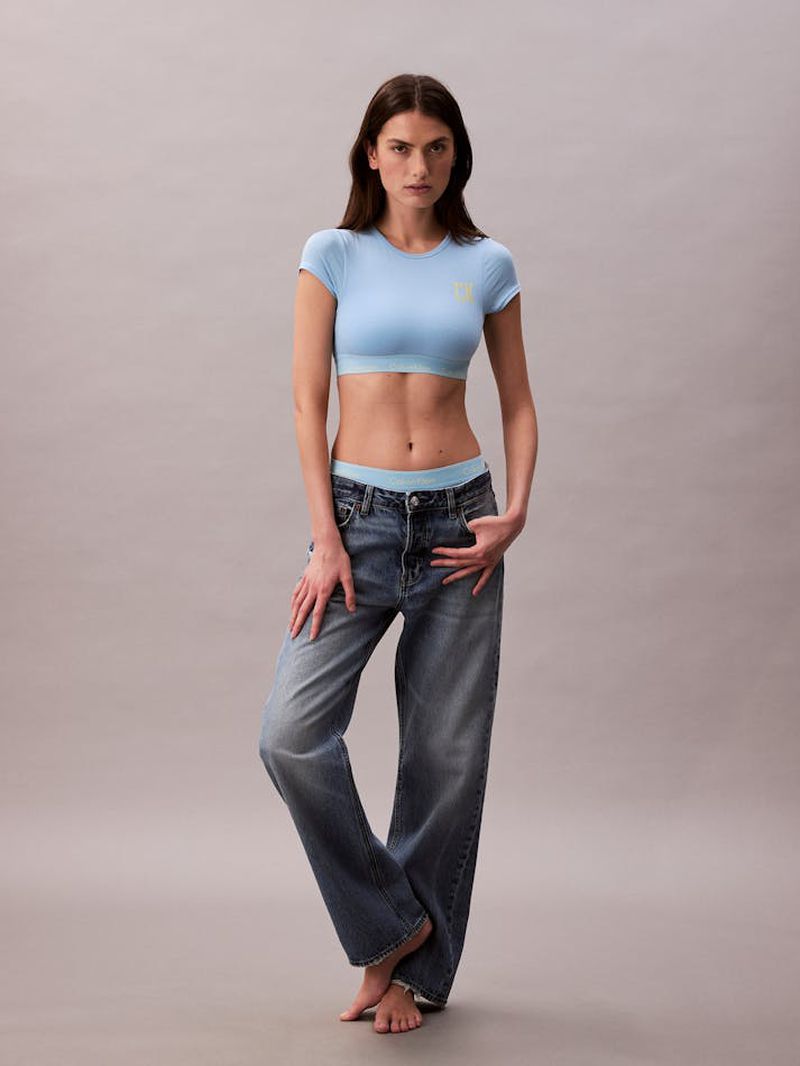 Full-length portrait of a young woman in a light blue top and blue jeans, posing in a studio setting with soft, diffused lighting.