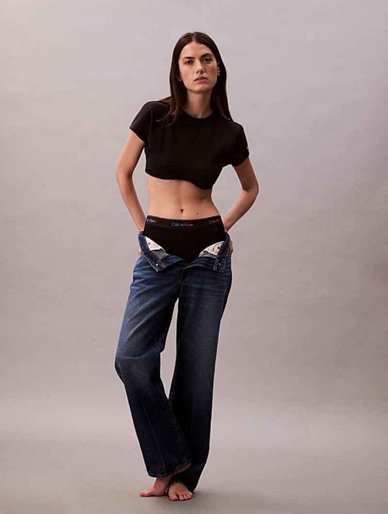 Model Mili Boskovic standing against a grey backdrop in a black crop top and unbuttoned denim jeans revealing black underwear.