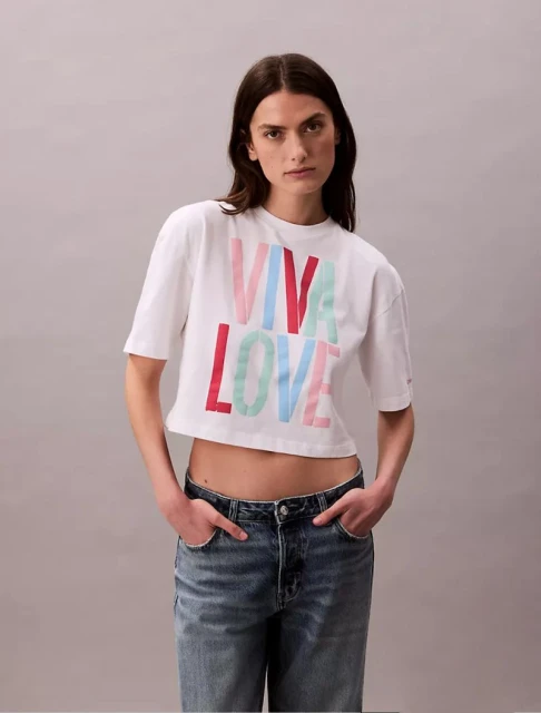 A young woman poses in a cropped t-shirt with the colorful text 'VIVA LOVE' and blue jeans against a neutral background in a studio shot.