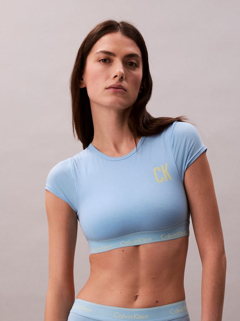 A woman poses indoors wearing a light blue Calvin Klein crop top and matching underwear, with a light grey background. Fashion shot.