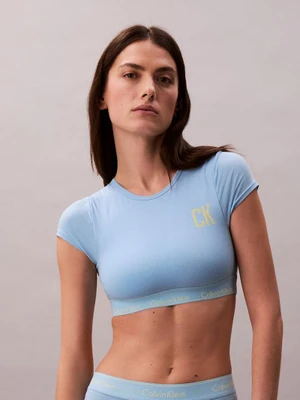 Model wearing a light blue cropped t-shirt with a yellow CK logo against a plain grey seamless studio background.