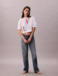 Model Mili Boskovic standing barefoot wearing a white Viva Love cropped t-shirt and blue denim jeans against a grey studio backdrop.