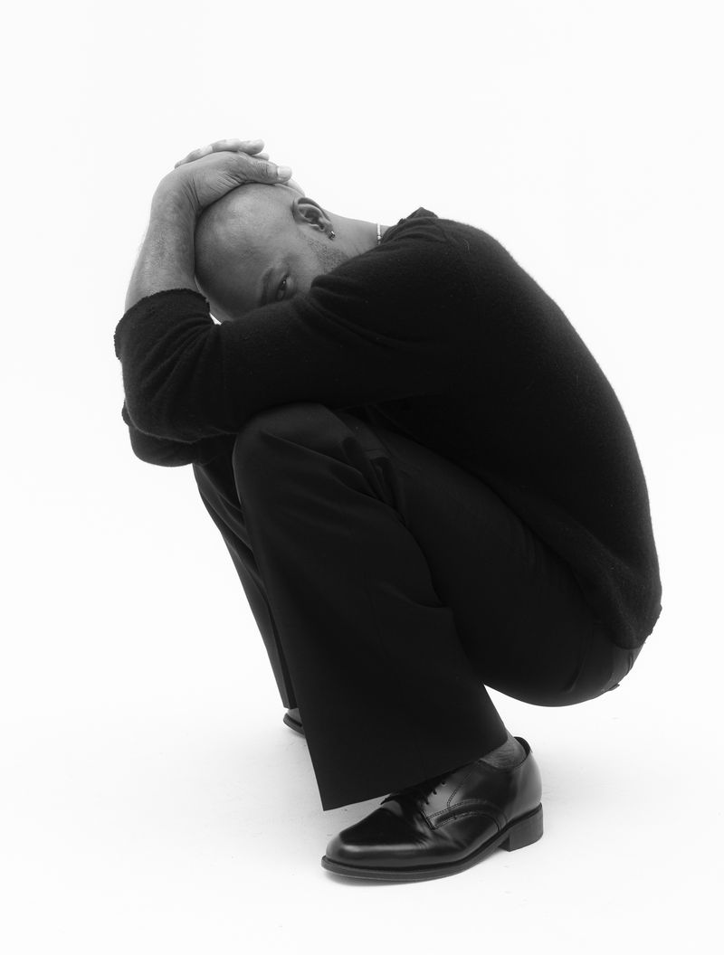 Monochrome portrait of a bald adult male in a crouching pose with his head resting on his hands, wearing dark clothing against white.
