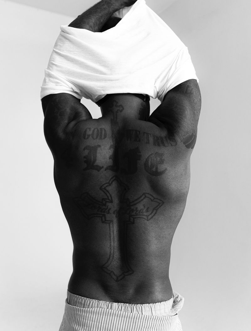 Black and white photo of a man's back with tattoos as he pulls a white t-shirt over his head in front of a plain background.