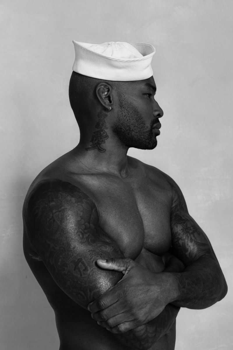 Monochrome portrait of muscular, tattooed adult male with sailor cap, arms crossed, eyes averted, against a textured neutral backdrop.