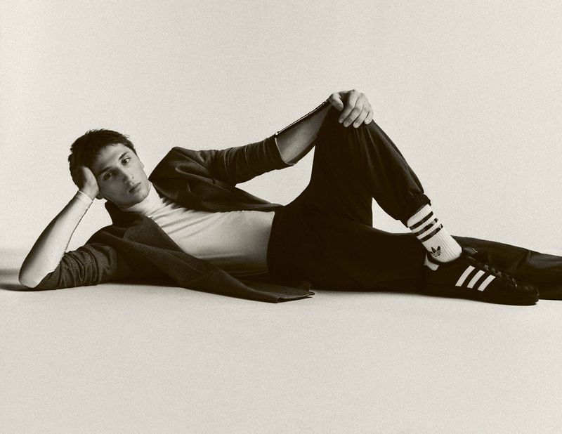 Monochrome portrait of a young adult male in casual fashion lying on a light backdrop in a studio setting, showcasing sneakers and relaxed pose.