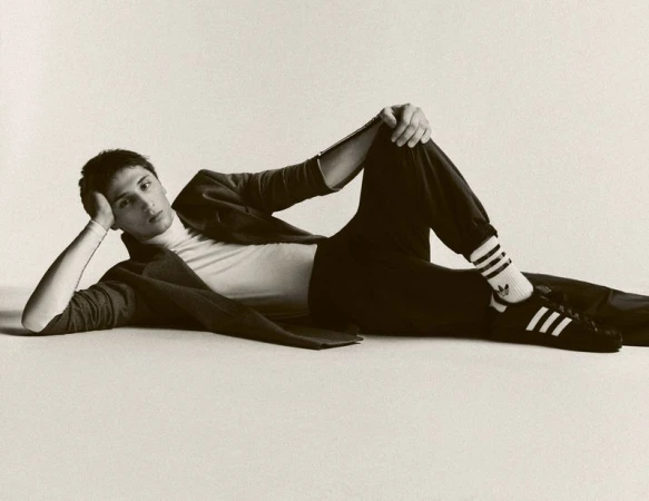 Monochrome portrait of a young adult male in casual fashion lying on a light backdrop in a studio setting, showcasing sneakers and relaxed pose.