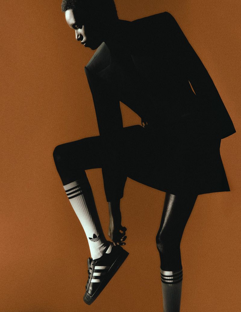 Dark-skinned adult model poses in black suit jacket, white socks with black stripes, and black sneakers against a brown background.