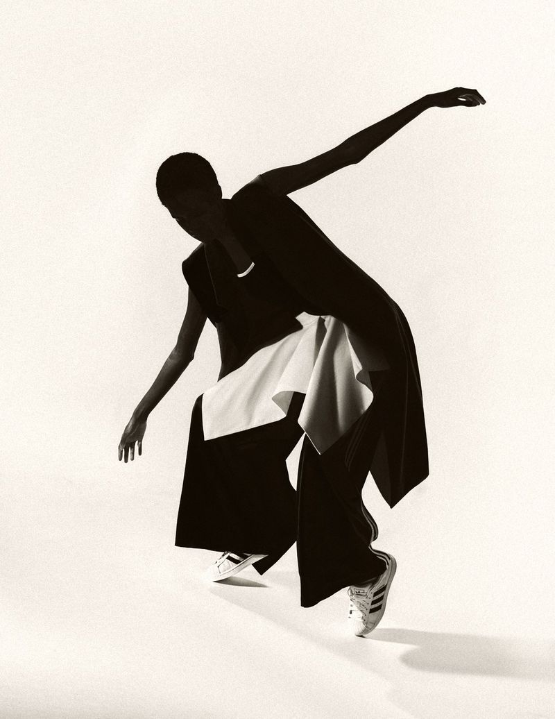 Woman in black outfit with a white scarf and Adidas sneakers strikes a dynamic pose against a bright white backdrop in a studio setting.