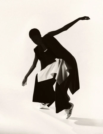 Woman in black outfit with a white scarf and Adidas sneakers strikes a dynamic pose against a bright white backdrop in a studio setting.