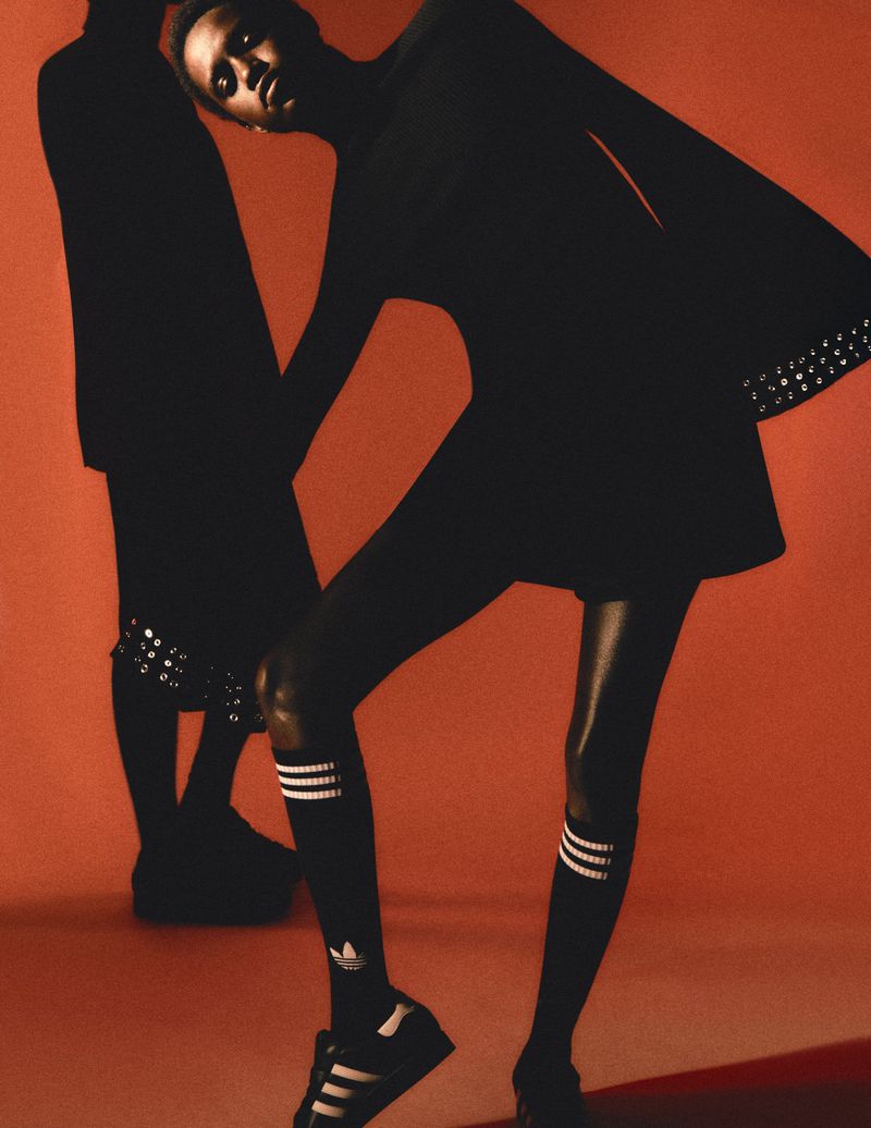 Two fashion models pose in black dresses, knee-high socks and sneakers against a terracotta background in a studio fashion shoot.