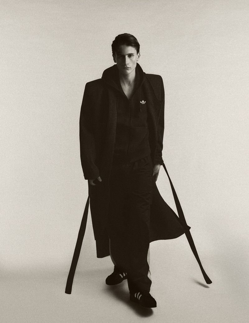 Full-length shot of a male fashion model posing in an all-black Adidas tracksuit and long black coat against a light neutral backdrop.