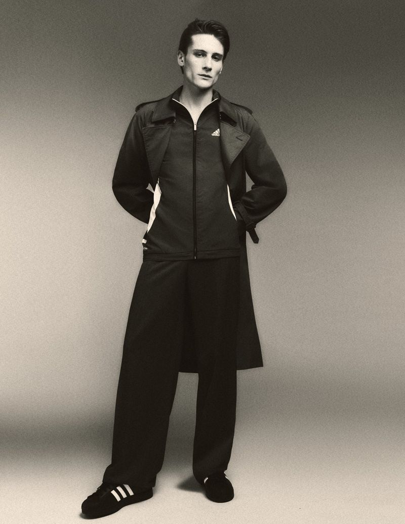 Monochrome portrait of a slender man wearing a dark Adidas tracksuit and long jacket in a studio setting against a grey backdrop.