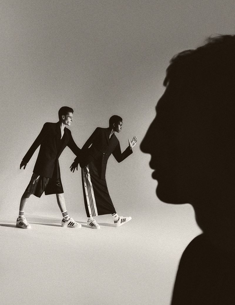Monochrome studio shot of two models in black suits and Adidas sneakers, one with a dark silhouette in the foreground.