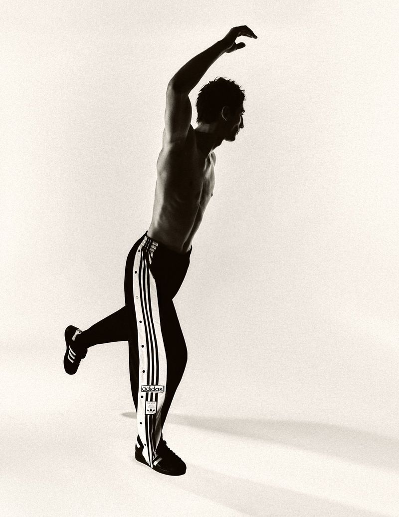 Monochromatic image of a muscular adult male posing in black and white track pants in a studio with a light background and dynamic lighting.